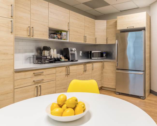A modern kitchen features light wooden cabinets, stainless steel appliances, and a round dining table with a bowl of lemons.