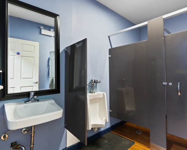 The image shows a bathroom with a wall-mounted sink, a urinal, and a shower stall separated by a partition, all set against blue walls.