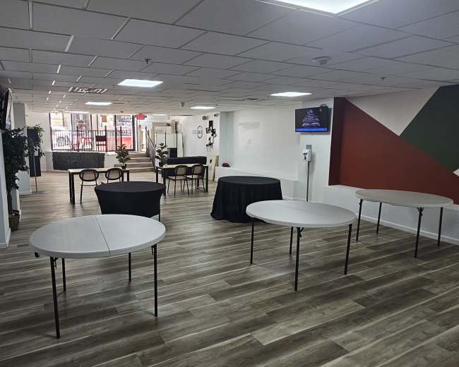 The image shows a spacious interior with several round and rectangular tables arranged, a seating area in the background, and a partially painted wall featuring geometric designs.