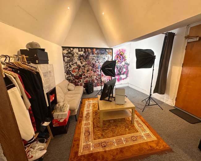 The image shows a small room with a couch, a coffee table, a television, photography lighting setups, and a wall covered in posters and artwork.