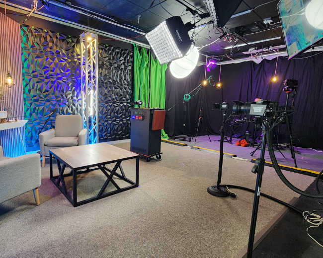 The image shows a television studio setup with two gray chairs, a coffee table, and various lighting and camera equipment arranged around a colorful backdrop.