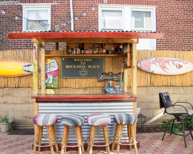A beach bar with a wooden counter and stools displays surfboards and a sign welcoming visitors to Rockaway Beach.