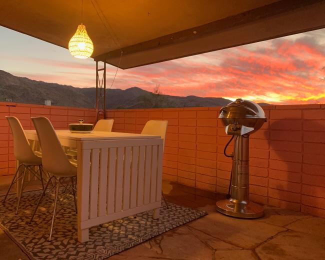 A outdoor dining area features a white table with four chairs and a grill, set against a backdrop of a vibrant sunset over the mountains.
