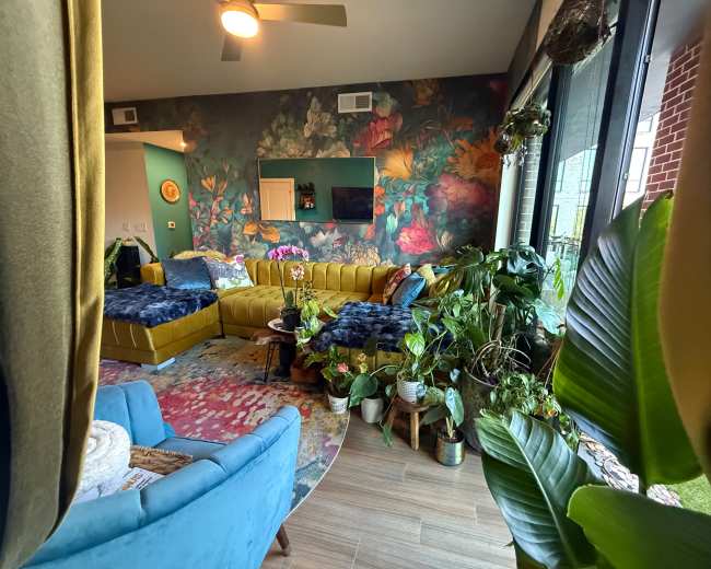 The image shows a vibrant living room featuring a large yellow sectional sofa, a decorative wall covered in floral wallpaper, and an assortment of indoor plants.
