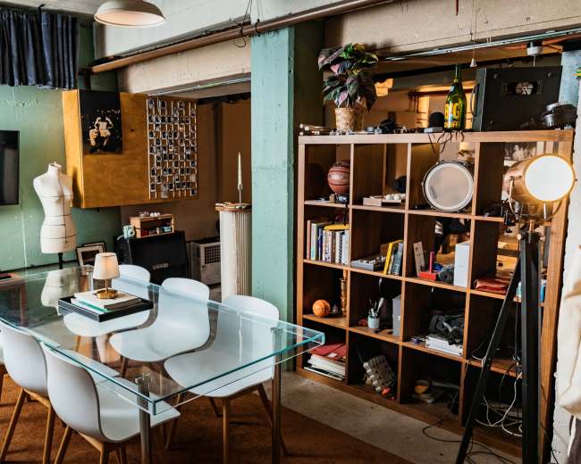 The image shows a modern dining area featuring a glass table surrounded by white chairs, with a bookshelf filled with various objects and decor in the background.