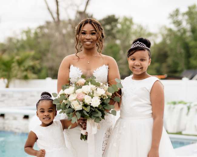 A bride stands by a pool, holding a bouquet of flowers, alongside two young girls in white dresses.