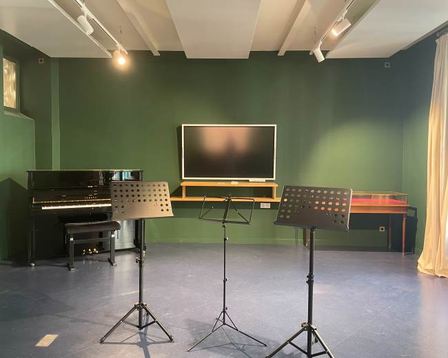 The image shows a music rehearsal space featuring a piano, three music stands, and a wall-mounted television against a green backdrop.