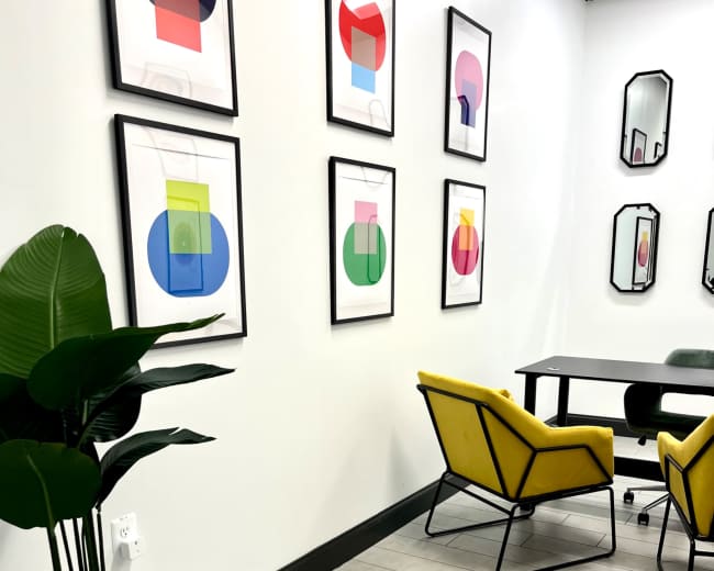 A modern office space with yellow chairs, a black table, a potted plant, and framed abstract art on the walls.