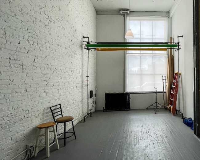 A spacious, empty studio features white brick walls, a large window, and a backdrop stand holding green and yellow fabric.