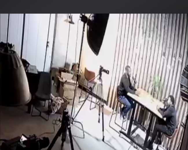 A camera captures two people sitting at a table in an office setting, surrounded by equipment and furniture.