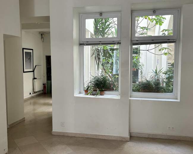 The image shows an interior space with white walls, two large windows allowing natural light, and a tiled floor.