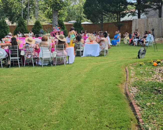 A gathering of people is set up in a backyard, with rows of chairs facing a decorated table, under a blue sky and surrounded by trees.