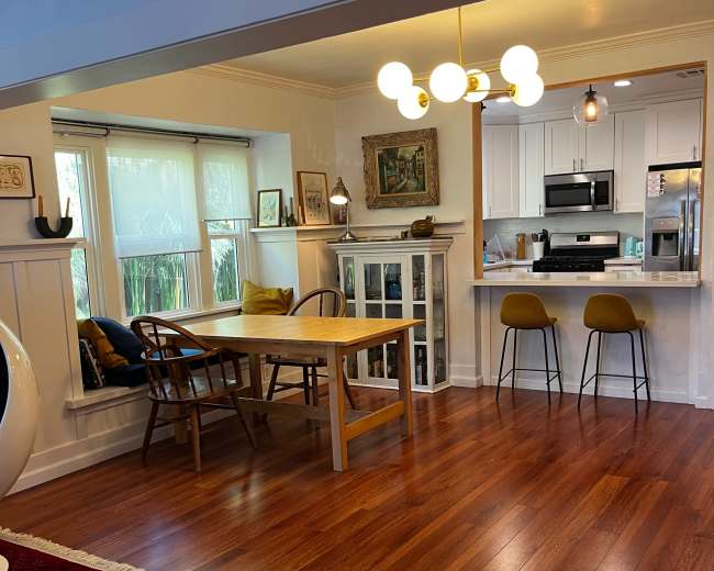 The image shows a bright, open living space featuring a dining table and chairs, a kitchen with modern appliances, and large windows allowing natural light to illuminate the wooden floors.