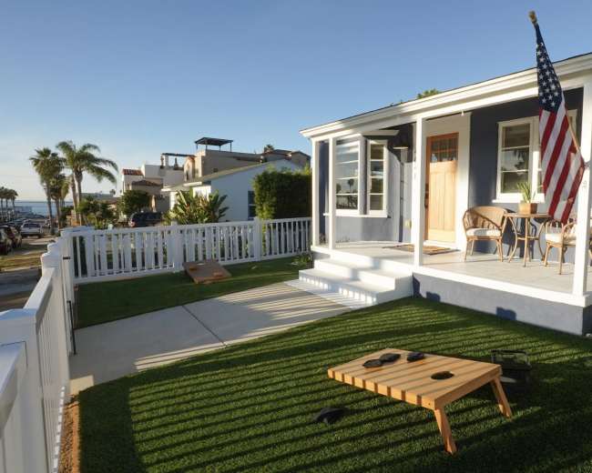 A blue house with a white porch displays an American flag, while two chairs sit on a grassy yard next to a cornhole game.