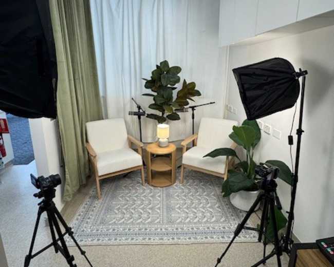 The image shows a small studio setup with two chairs, a low table, two camera tripods with cameras, and decorative plants against a backdrop of curtains.