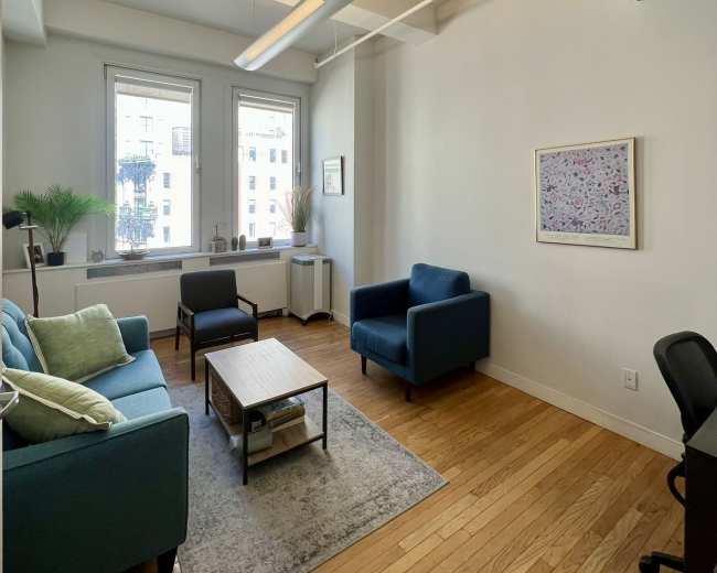 The image shows a bright office space with two blue couches, a coffee table, and a desk, featuring large windows that let in natural light.