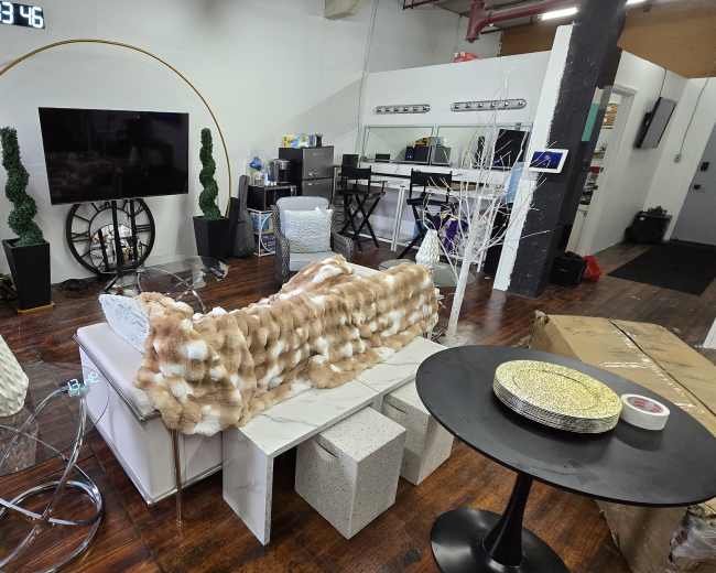 A modern interior space features a white couch with a fuzzy blanket, a glass coffee table, decorative plants, and a large television mounted on the wall.