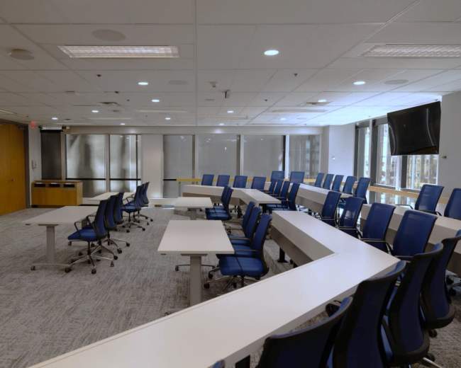 The image shows a modern conference room with a circular arrangement of white tables and blue chairs, equipped with large windows and two screens on the walls.