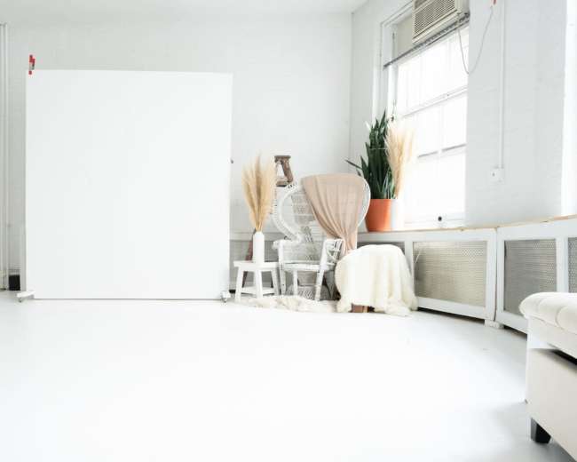The image shows a bright, minimalistic studio space featuring a large white backdrop, a chair, potted plants, and a cozy throw on a bench.