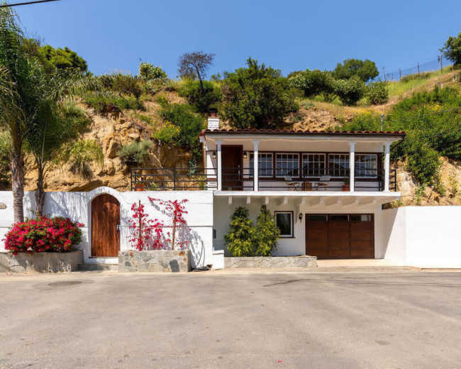 A two-story white house with a brown tiled roof and a garage is situated on a sloped lot surrounded by greenery.