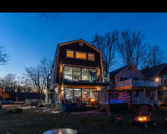 The image shows a modern two-story house with large windows and an outdoor seating area, set against a twilight sky.