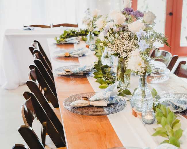 A long wooden table is elegantly set with floral centerpieces, dinnerware, and greenery, surrounded by black chairs in a decorated indoor venue.