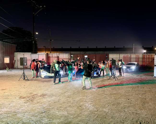 A group of people is gathered around two cars in an outdoor setting at night, with colorful lights illuminating the scene.