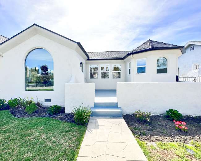 The image shows a white stucco house with a curved front wall, large windows, and a stone pathway leading to the entrance.