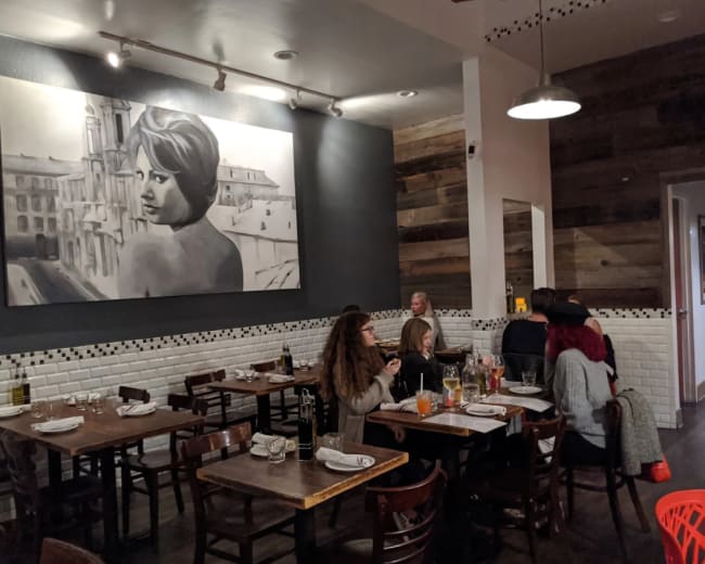 A modern restaurant features wooden tables and chairs, with a large black and white mural of a woman on the wall and patrons sitting at tables.
