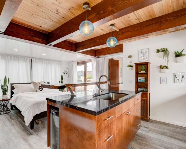 A modern studio apartment with wooden beams, a kitchen island, and a bed area separated by sheer curtains.