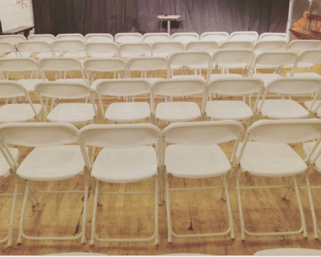 The image shows an empty auditorium setup with rows of white folding chairs facing a small stage covered by a black curtain.