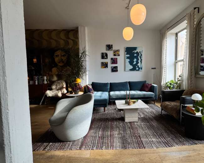 The image shows a modern living room with a teal sofa, a light-colored coffee table, and various plants, framed artwork, and warm lighting.
