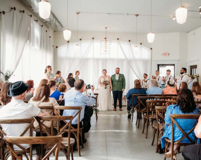 A wedding ceremony is taking place in a well-lit venue, with guests seated on wooden chairs and the bridal party standing at the front.