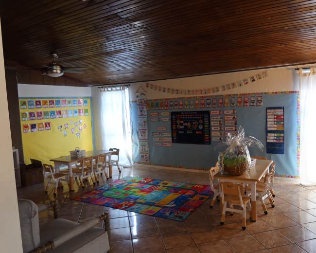 A brightly lit classroom features colorful educational posters on the walls, a large rug on the floor, and several small tables and chairs arranged for students.