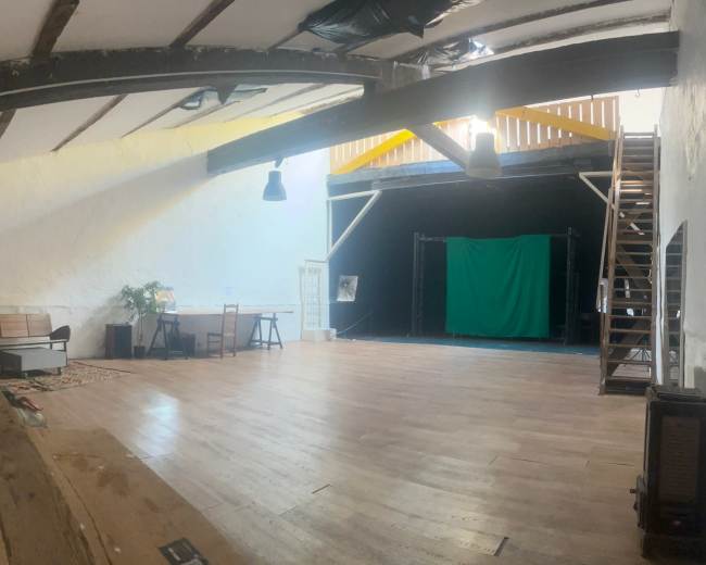 The image shows a spacious, well-lit studio with wooden beams, a large wooden floor, and a green screen setup in one corner.