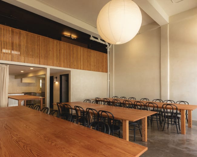 The image shows a spacious dining area with long wooden tables and black chairs, illuminated by a large round pendant light.