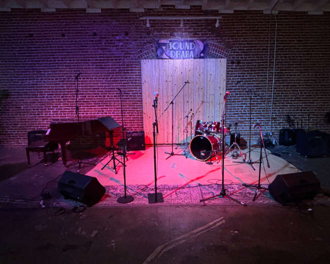 The image shows an empty stage setup with musical instruments, including a piano and a drum set, surrounded by microphone stands and speakers, against a brick wall backdrop.