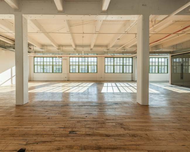 The image depicts a spacious, empty room with wooden floors and large windows allowing natural light to illuminate the space.