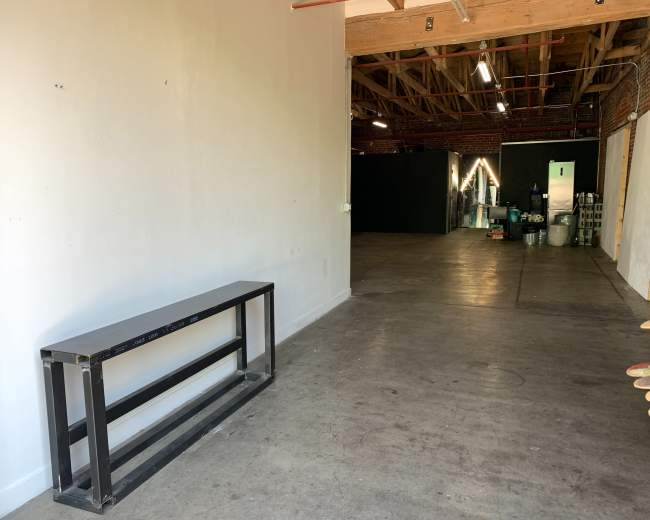 The image shows a spacious, empty room with a metal table against a white wall and a dark area visible in the background.