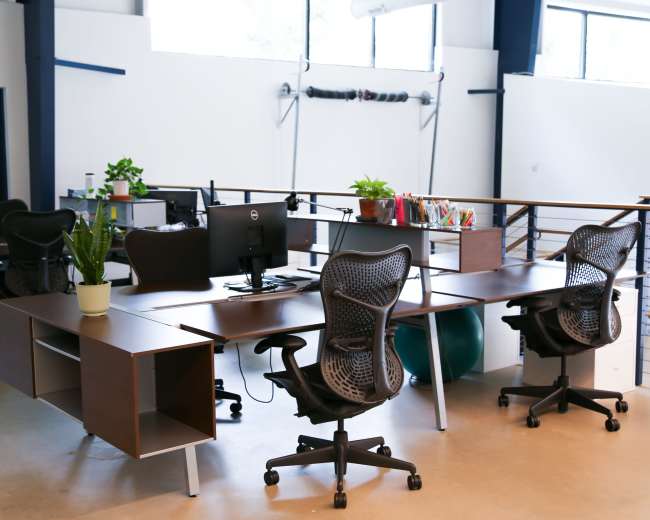 The image shows a modern office space featuring several desks with ergonomic chairs, plants, and computer equipment arranged in an open layout.