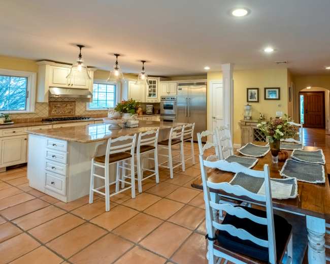 The image shows a spacious kitchen featuring a central island with bar seating, a wooden dining table, and tile flooring.