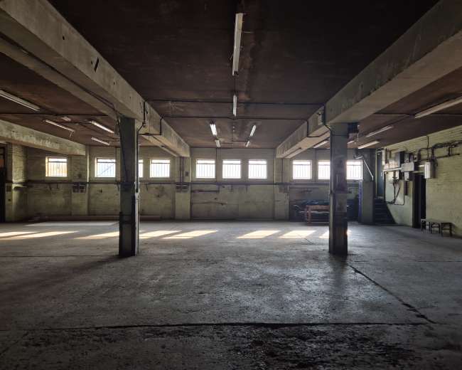 The image shows an empty industrial warehouse with concrete floors and large windows allowing sunlight to stream in.