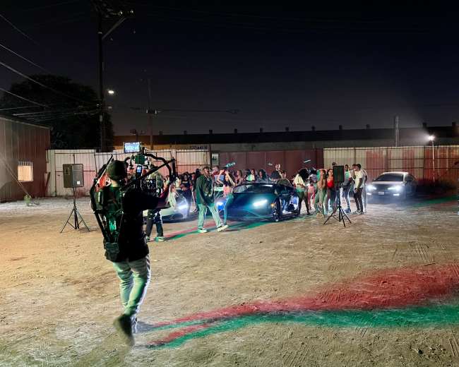 A film crew shoots a scene featuring a group of people near flashy cars at night in an outdoor setting.