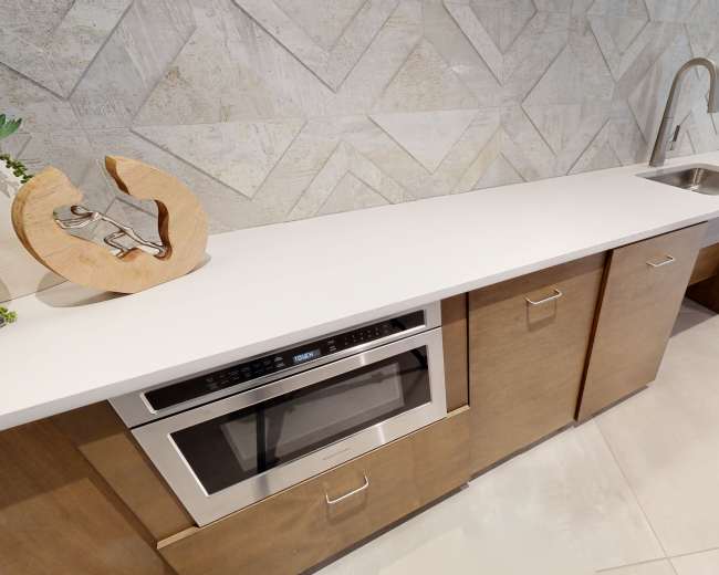 A modern kitchen countertop featuring a sink, built-in microwave, and wooden cabinetry against a textured wall.