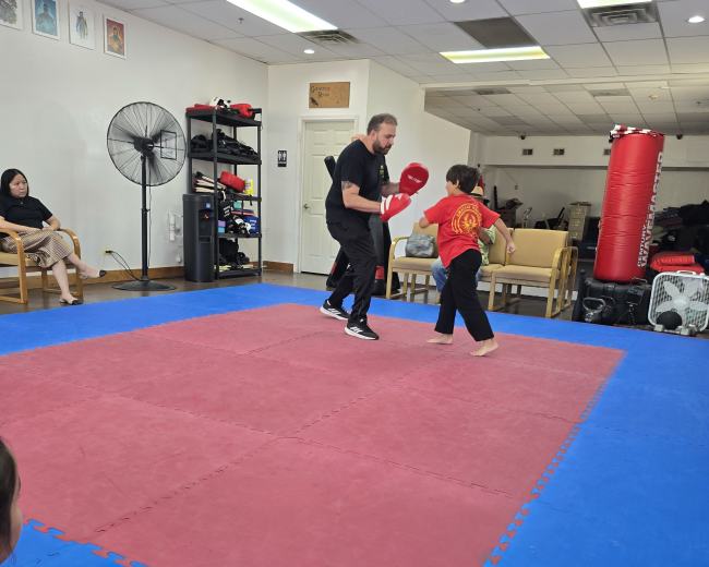 A trainer demonstrates boxing techniques to a young student in a martial arts studio while others observe.