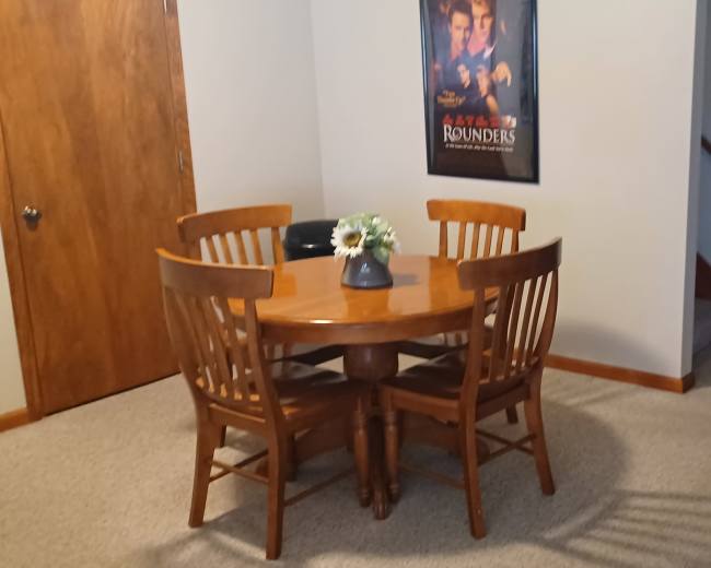 A wooden dining table with four chairs sits near a wall adorned with a movie poster, adjacent to a hallway leading to another room.