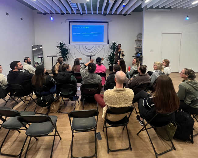 A group of people sits in chairs facing a speaker in a well-lit, modern room with a presentation screen.