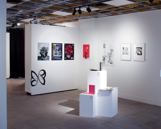 The image shows an art gallery interior with white walls displaying various framed artworks and a sculptural display stand in the center.