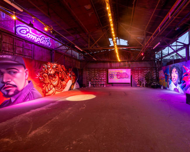 The image shows a spacious indoor venue with vibrant graffiti murals on the walls and colorful lighting illuminating the area.