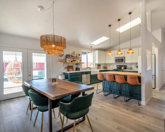 The image shows a modern kitchen and dining area featuring a large table with green chairs, stylish pendant lighting, and a color palette of teal and wood tones.
